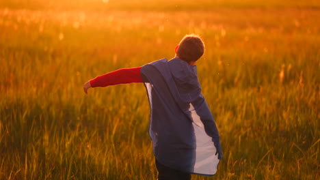 A-boy-in-a-suit-and-a-superhero-mask-with-a-red-cloak-runs-across-the-field-at-sunset-on-the-grass-dreaming-and-imagining-himself-a-hero.